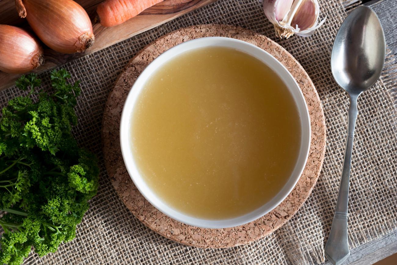 Chicken bone broth served in a soup bowl, with onions, carrots, parsley and garlic in the background, top view