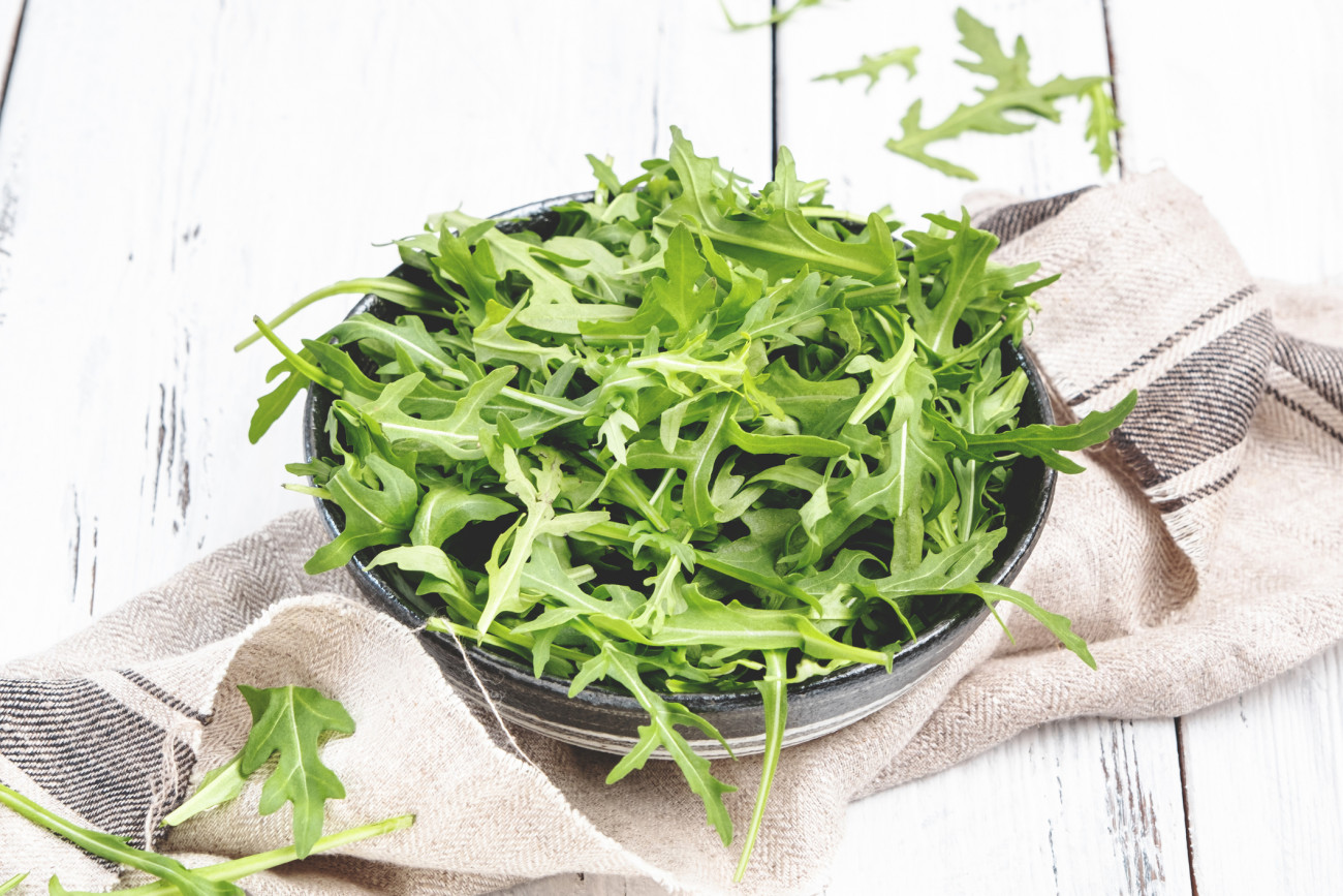 Fresh green arugula in rustic ceramic bowl on white kitchen table background, top view, copy space. Spring spicy herb for healthy eating
